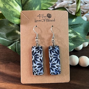 May include: A pair of rectangular earrings with a floral pattern in navy blue and white. The earrings hang from silver-colored hooks and are displayed on a brown card with the text "4 The Love Of Wood".