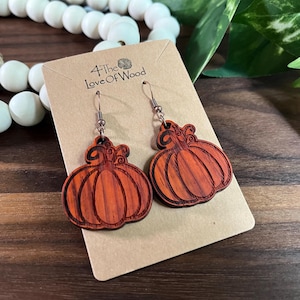 May include: A pair of orange pumpkin-shaped wooden earrings with dark brown engraved lines, hanging from metal hooks on a tan cardboard display card labeled 4 The Love Of Wood. The earrings feature a natural wood grain texture and curved stems.