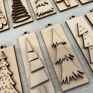 May include: A collection of rectangular light-colored wooden ornaments, each featuring a unique laser-cut Christmas tree design. The ornaments include various tree shapes, some with dotted patterns, others with layered geometric segments.