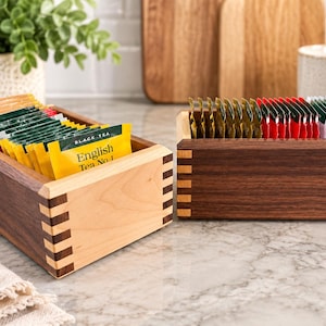 May include: Two rectangular wooden storage boxes with box joint corners. The left box features light-colored wood sides and dark wood ends, while the right box is primarily dark wood. Both are filled with various tea bags, including one labeled English Tea No. 1.
