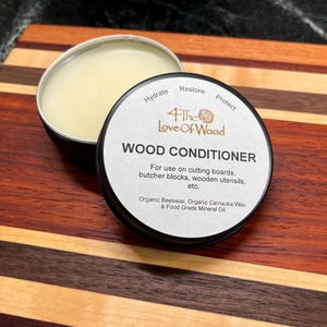 May include: A round tin of wood conditioner with a black lid. The label says "4 The Love Of Wood" and "Wood Conditioner". It also says "For use on cutting boards, butcher blocks, wooden utensils, etc." and "Organic Beeswax, Organic Carnauba Wax & Food Grade Mineral Oil".