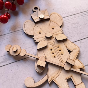 May include: Wooden Christmas ornament in the shape of a mouse soldier wearing a crown and holding a staff. The ornament is made of light-colored wood and has intricate cut-out details. Red berries are visible in the background.