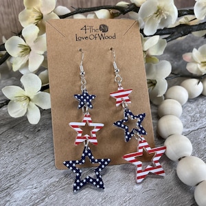 May include: Dangling star earrings with patriotic red, white, and blue designs. The earrings feature a layered star design with the American flag pattern. The earrings are hanging on a brown card with the text "4 The Love Of Wood".