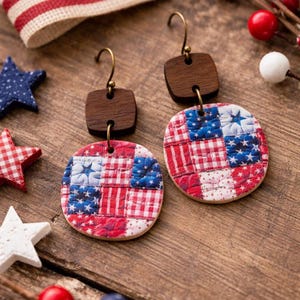 May include: A pair of patriotic earrings with a wooden top and a circular, patchwork design. The earrings feature red, white, and blue fabric squares, with stars and stripes patterns. The earrings are displayed on a wooden surface.