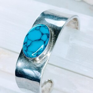 May include: A silver cuff bracelet with a turquoise stone set in the centre. The stone has a blue and green colour and a marbled pattern.