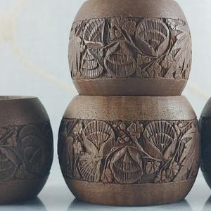 May include: Four brown wooden candle holders with carved floral designs. The candle holders are cylindrical with a rounded top and bottom. The carvings feature detailed leaves and flowers. The candle holders are stacked in a pyramid shape.