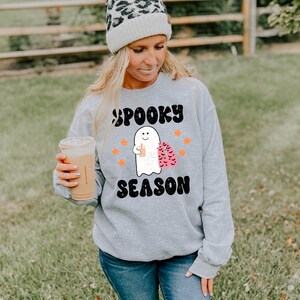 May include: A grey sweatshirt with a cartoon ghost holding a drink and the text "Spooky Season". The ghost is wearing a pink cape with black bats. The sweatshirt has a white pom-pom beanie on top.
