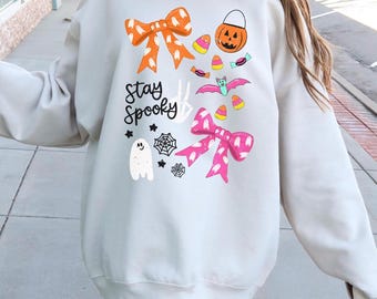 Może przedstawiać: Kremowa bluza z motywem Halloween. Projekt zawiera pomarańczowe i różowe kokardy, dynię-latarnię, cukierki kukurydziane, nietoperza, ducha i napis "Stay Spooky".