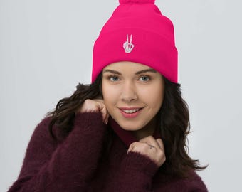 Może przedstawiać: Żywa różowa czapka z pasującym pomponem. Czapka ma białą haftowaną dłoń szkieletu z znakiem pokoju. Stylowy dodatek na zimę.