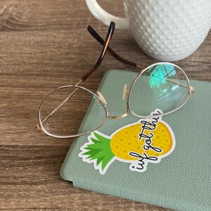 May include: Gold-rimmed round glasses with clear lenses resting on a green leather notebook with a pineapple sticker that says "I've got this".