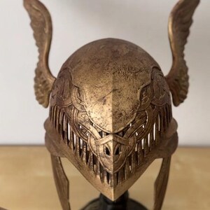 K&ouml;nnte beinhalten: Ein goldfarbener Helm mit Fl&uuml;geln an den Seiten. Der Helm hat komplizierte Details und eine strukturierte Oberfl&auml;che.