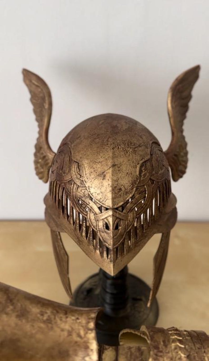 K&ouml;nnte beinhalten: Ein goldfarbener Helm mit Fl&uuml;geln an den Seiten. Der Helm hat komplizierte Details und eine strukturierte Oberfl&auml;che.