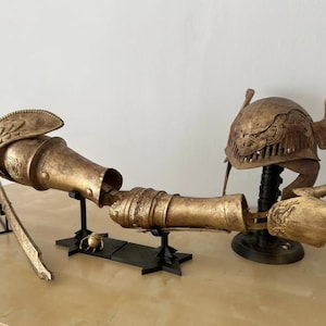 K&ouml;nnte beinhalten: Eine goldfarbene, artikulierte Arm-Skulptur mit Helm und Fl&uuml;geln. Der Arm steht auf einem schwarzen St&auml;nder und der Helm auf einem separaten St&auml;nder. Der Arm ist aus Metall und hat ein detailliertes Design.