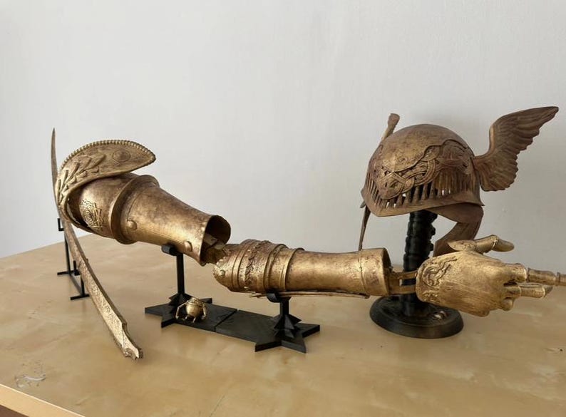 K&ouml;nnte beinhalten: Eine goldfarbene, artikulierte Arm-Skulptur mit Helm und Fl&uuml;geln. Der Arm steht auf einem schwarzen St&auml;nder und der Helm auf einem separaten St&auml;nder. Der Arm ist aus Metall und hat ein detailliertes Design.