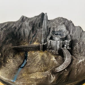 Peut inclure: Modèle imprimé en 3D d'un château fantastique avec une haute tour, entouré d'un mur de pierre et d'un chemin sinueux. Le château est situé sur une base rocheuse avec une petite rivière qui le traverse.