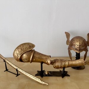 K&ouml;nnte beinhalten: Ein goldfarbenes Set an Fantasy-R&uuml;stungen, bestehend aus einem Helm mit Fl&uuml;geln, einem Schwert und einem Unterarmschutz. Die Teile werden auf schwarzen St&auml;ndern pr&auml;sentiert.