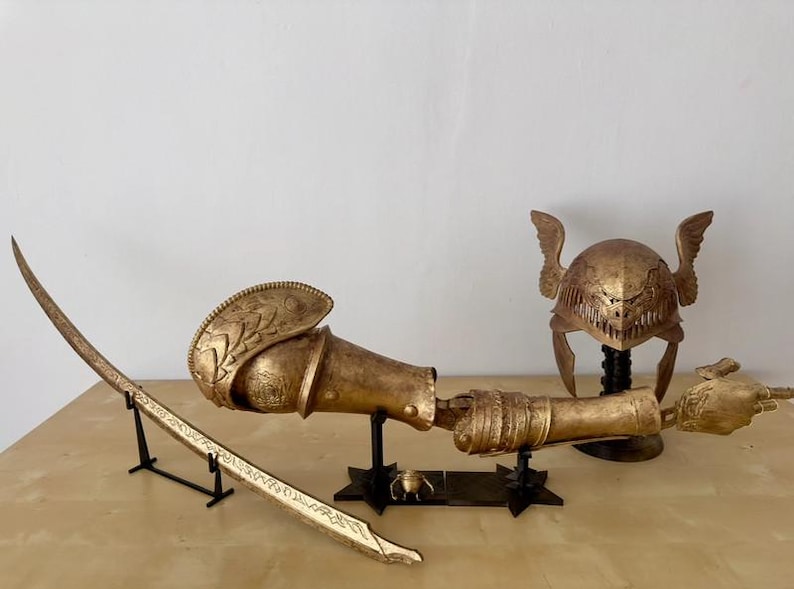 K&ouml;nnte beinhalten: Ein goldfarbenes Set an Fantasy-R&uuml;stungen, bestehend aus einem Helm mit Fl&uuml;geln, einem Schwert und einem Unterarmschutz. Die Teile werden auf schwarzen St&auml;ndern pr&auml;sentiert.