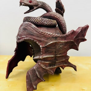 Könnte beinhalten: Ein rot-brauner Helm mit einer darauf modellierten Schlange. Der Helm hat an den Seiten ein flügelartiges Design.