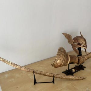 K&ouml;nnte beinhalten: Ein goldfarbenes Schwert, Helm und Armr&uuml;stung auf einem schwarzen St&auml;nder. Das Schwert ist gebogen und mit komplizierten Details verziert. Der Helm hat Fl&uuml;gel und ist mit einem Muster verziert. Die Armr&uuml;stung ist ebenfalls mit einem Muster verziert.