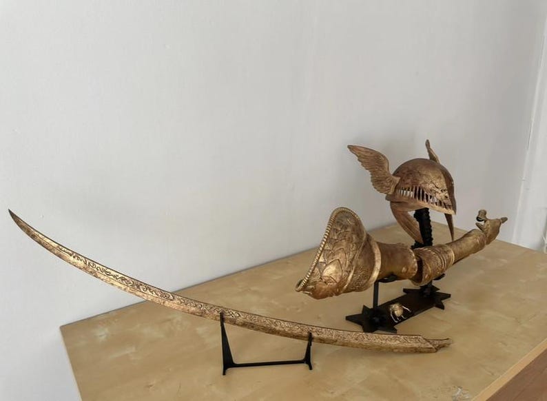 K&ouml;nnte beinhalten: Ein goldfarbenes Schwert, Helm und Armr&uuml;stung auf einem schwarzen St&auml;nder. Das Schwert ist gebogen und mit komplizierten Details verziert. Der Helm hat Fl&uuml;gel und ist mit einem Muster verziert. Die Armr&uuml;stung ist ebenfalls mit einem Muster verziert.