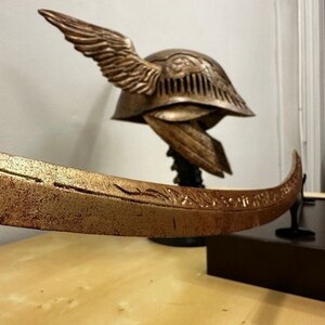 K&ouml;nnte beinhalten: Ein goldfarbener Helm mit Fl&uuml;geln und filigranen Details sowie ein passendes Schwert mit einer gebogenen Klinge. Helm und Schwert werden auf einem Holzst&auml;nder pr&auml;sentiert.