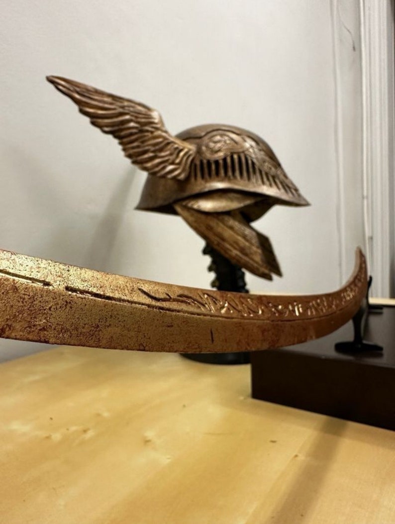 K&ouml;nnte beinhalten: Ein goldfarbener Helm mit Fl&uuml;geln und filigranen Details sowie ein passendes Schwert mit einer gebogenen Klinge. Helm und Schwert werden auf einem Holzst&auml;nder pr&auml;sentiert.