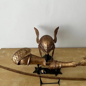 K&ouml;nnte beinhalten: Ein goldfarbener, detaillierter Kriegerhelm mit Fl&uuml;geln, einem passenden Arm und einer Hand sowie einem Schwert auf einem schwarzen St&auml;nder.
