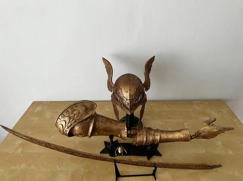 K&ouml;nnte beinhalten: Ein goldfarbener, detaillierter Kriegerhelm mit Fl&uuml;geln, einem passenden Arm und einer Hand sowie einem Schwert auf einem schwarzen St&auml;nder.