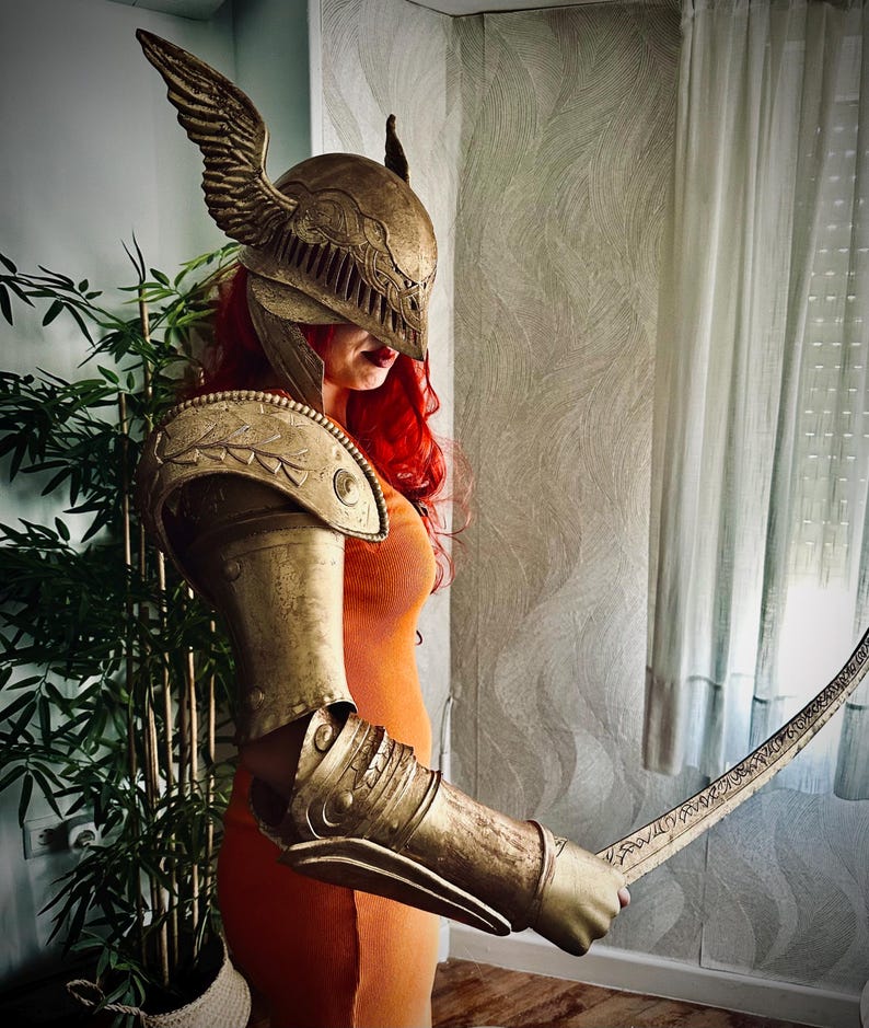 K&ouml;nnte beinhalten: Eine Frau tr&auml;gt einen goldenen Helm mit Fl&uuml;geln und eine passende R&uuml;stung und h&auml;lt ein Schwert mit kunstvollen Gravuren.