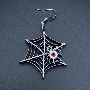 May include: Silver spider web earring with a red and white spider in the center.