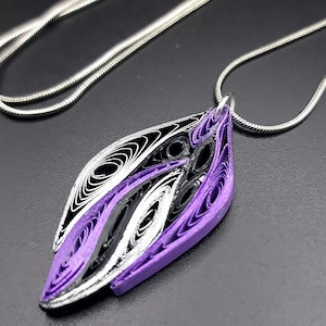 Pode incluir: Um colar de corrente prateada com um pingente de papel quilled roxo, preto e prateado. O pingente tem a forma de uma lágrima e apresenta um padrão em espiral.