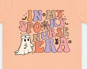 Może przedstawiać: Różowy t-shirt z napisem "In My Spooky Nurse Era" w retro stylu. Tekst jest w odcieniach różowego, fioletowego i pomarańczowego. Po lewej stronie tekstu znajduje się kreskówkowy duch z czapką pielęgniarki i strzykawką.