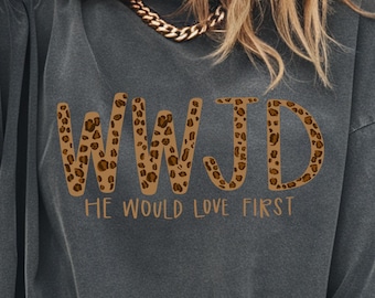 May include: Dark gray t-shirt with the text "WWJD" in a leopard print pattern and the words "HE WOULD LOVE FIRST" in a brown font. The shirt has short sleeves and a relaxed fit.