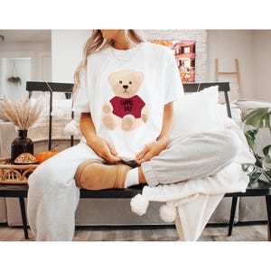 May include: White t-shirt featuring a cartoon teddy bear wearing a maroon shirt with the text "GAME DAY". The bear has a tan body, black eyes, and a black nose. The shirt is a casual, comfortable style.