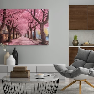 May include: A canvas print of a pink cherry blossom tree lined pathway. The pathway is lined with trees on both sides and leads to a building in the distance. The print is hanging on a wall in a living room setting with a grey armchair and a coffee table.