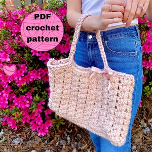 May include: A pink crocheted tote bag with a pink ribbon bow. The bag is being held by a person wearing blue jeans. The text "PDF crochet pattern" is visible in a pink circle.