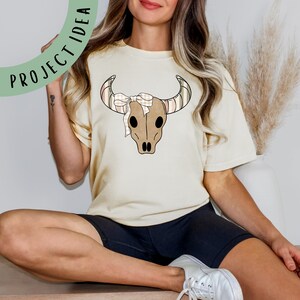 Puede incluir: Una camiseta de color crema con un gr&aacute;fico de cr&aacute;neo de vaca marr&oacute;n y blanco con un lazo de cuadros rosa y blanco.