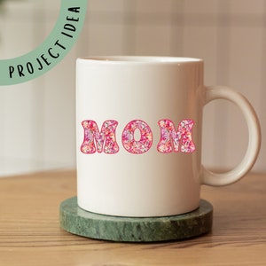 May include: White ceramic mug with a pink floral design spelling out the word "MOM".