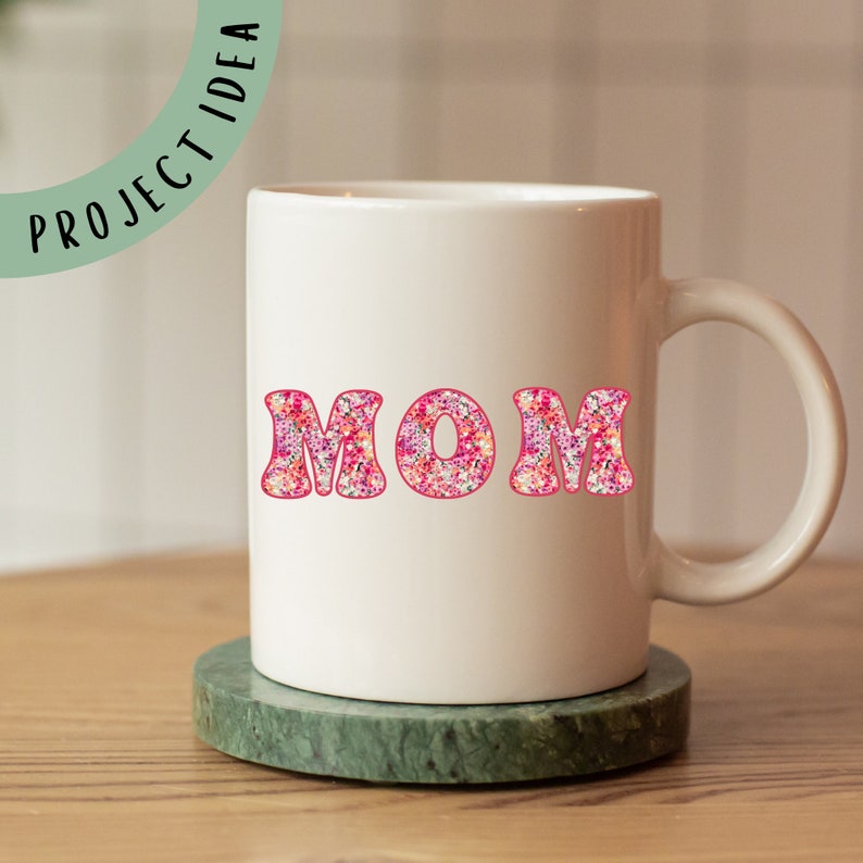 May include: White ceramic mug with a pink floral design spelling out the word "MOM".