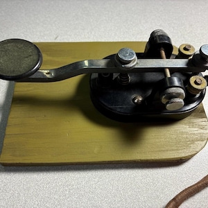 May include: A vintage telegraph key mounted on a rectangular olive green wooden base. The device features a black plastic oval housing, a silver metal lever with a circular black knob, and several silver adjustment screws with a brown fabric-covered wire.