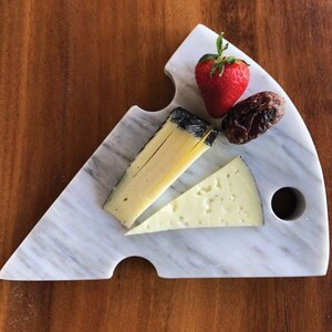 May include: A marble cheese board with a semi-circular shape, featuring a strawberry, a date, and two types of cheese. The board has a gray and white veined pattern. The cheese is yellow and the strawberry is red.