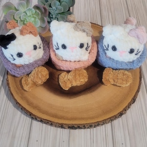 May include: Three handmade plush cat toys with different colored bows and collars. The cats are white with black eyes and pink noses. Each cat has a different colored collar: purple, pink, and blue. The toys are on a wooden slice.