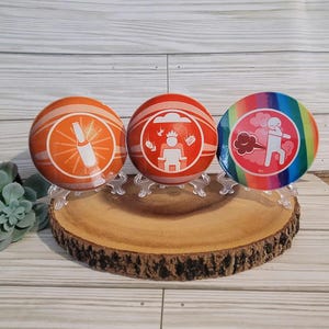 May include: Three round coasters with different designs. The first coaster is orange with a white outline and a white bottle with a sunburst behind it. The second coaster is orange with a white outline and a white stick figure wearing a crown sitting on a throne with a cloud above it. The third coaster is rainbow with a white outline and a white stick figure with a cloud of gas coming out of its rear end.