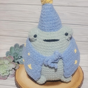 May include: A handmade crochet frog plushie wearing a light blue wizard hat and cape with yellow star accents. The frog is light green and gray, with black eyes and a stitched mouth. It sits on a wooden slice.