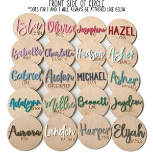 K&ouml;nnte beinhalten: Eine Sammlung von Holzscheiben mit Namen in verschiedenen Farben. Die Namen sind in Schreibschrift gehalten und umfassen "Isla", "Olivia", "Josephine", "Hazel", "Isabella", "Charlotte", "Hudson", "Asher", "Gabriel", "Arden", "Michael", "Asher", "Adalynn", "Millie", "Bennett", "Jayden", "Aurora", "Landon", "Harper" und "Elyah."
