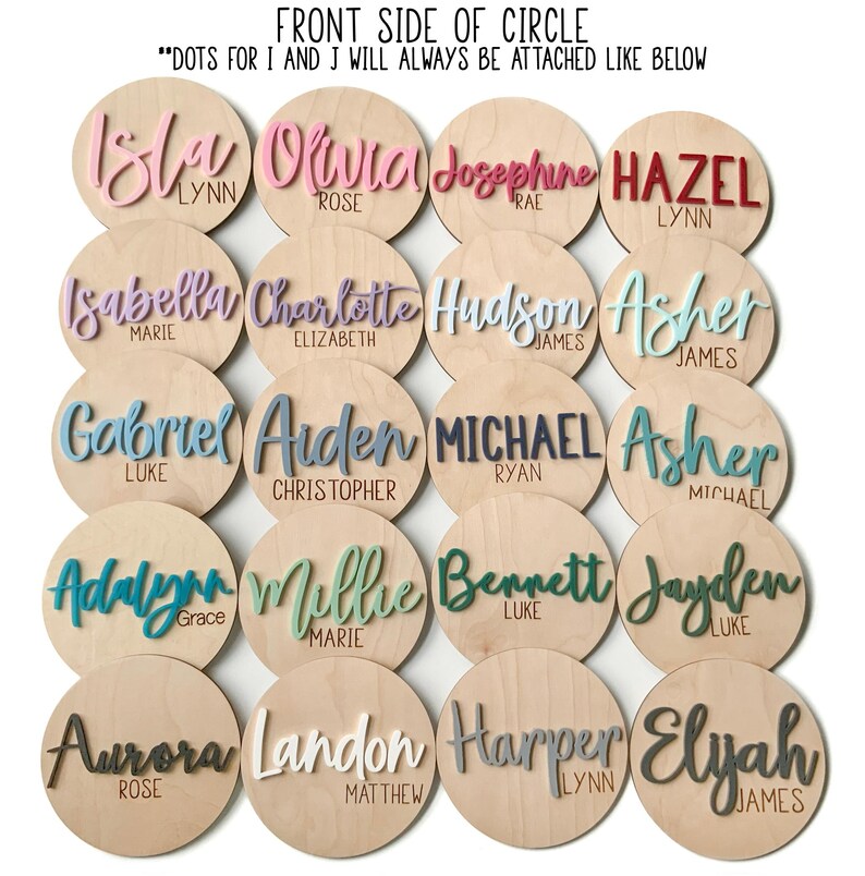 K&ouml;nnte beinhalten: Eine Sammlung von Holzscheiben mit Namen in verschiedenen Farben. Die Namen sind in Schreibschrift gehalten und umfassen "Isla", "Olivia", "Josephine", "Hazel", "Isabella", "Charlotte", "Hudson", "Asher", "Gabriel", "Arden", "Michael", "Asher", "Adalynn", "Millie", "Bennett", "Jayden", "Aurora", "Landon", "Harper" und "Elyah."