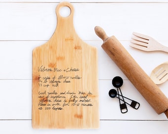 May include: A bamboo cutting board with a handle, featuring handwritten recipe text for Velveeta Mac + Cheese. A wooden rolling pin, measuring spoons, and wooden utensils are arranged nearby, suggesting a kitchen theme.