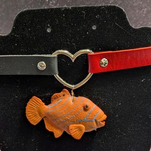 Pode incluir: Um colar de couro preto e vermelho com um pingente de coração prateado e um pequeno peixe laranja.
