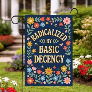 Op de afbeelding: Een marineblauwe tuinvaandel met de woorden "RADICALIZED BY BASIC DECENCY" in een gouden lettertype, omringd door kleurrijke bloemen, bijen en vlinders. De vlag hangt aan een zwarte metalen standaard in een tuin.