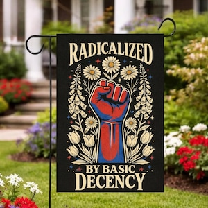 Pode incluir: Bandeira de jardim preta com o texto "RADICALIZED BY BASIC DECENCY" em uma fonte decorativa. A bandeira apresenta um punho vermelho e azul, rodeado por flores brancas e folhagem verde. A bandeira está pendurada em um suporte de metal preto.